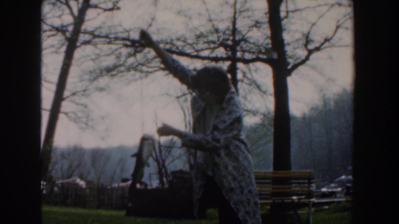In Novi, Michigan in 1961, a woman in glasses and a patterned coat raises a fish beside bare trees and a park bench.