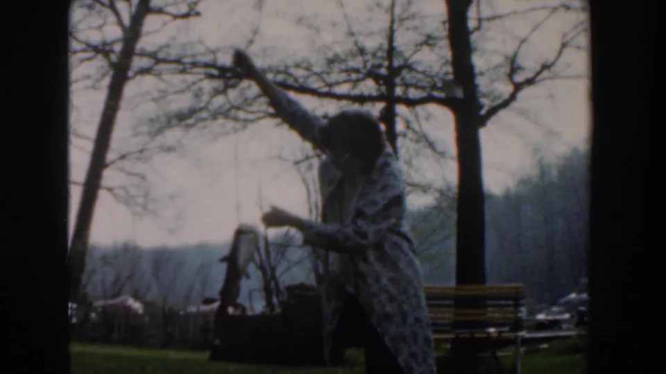 In Novi, Michigan in 1961, a woman in glasses and a patterned coat raises a fish beside bare trees and a park bench.