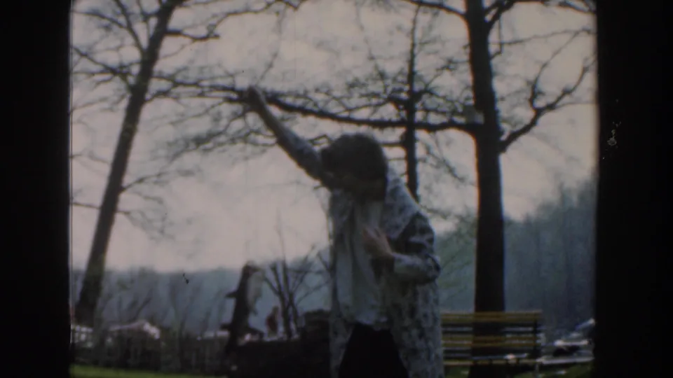 A 1961 home-movie frame shows the woman holding the fish high, partly shadowed, with a bench and bare park trees behind her.
