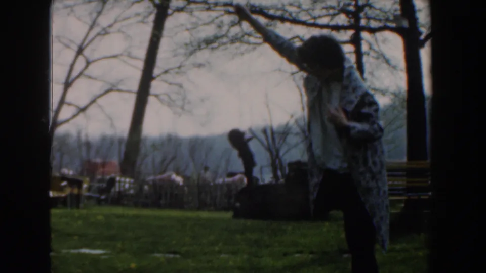 The woman stands in the grass with a fish raised overhead, with bare trees, a bench, and distant cars in the Novi park scene.