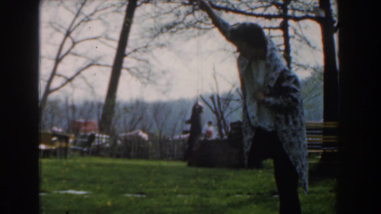 Person in a patterned blanket holds a fish aloft beside a bench and trees.