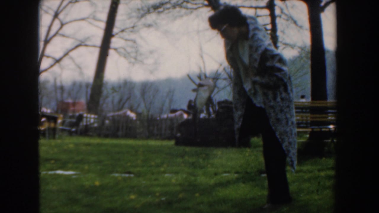 Woman bends slightly on a lawn beside fishing gear, with benches, trees, and distant buildings behind her.