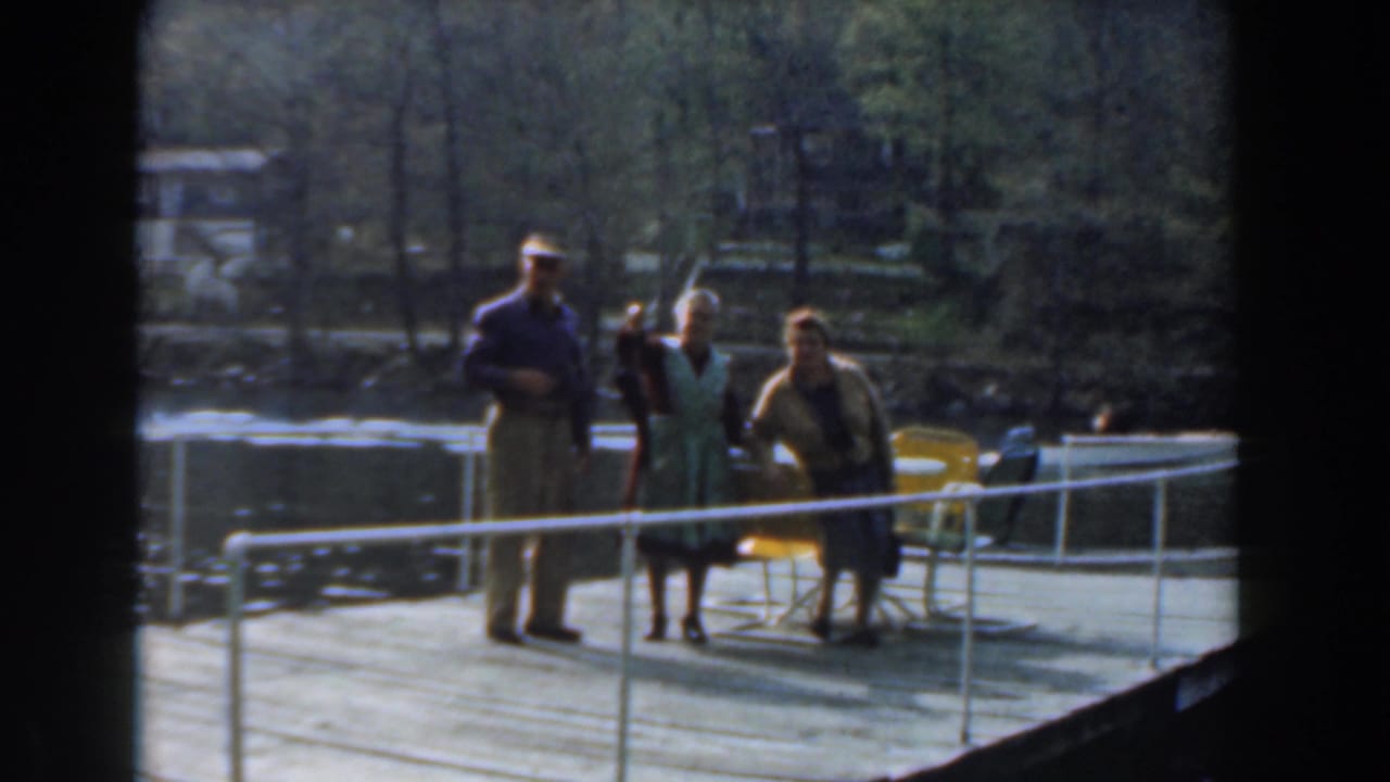 Three adults on a lakeside dock in Novi, Michigan, with yellow chairs, white railing, trees, and water visible in 1961 8mm footage.