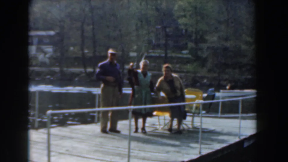Three adults on a lakeside dock in Novi, Michigan, with yellow chairs, white railing, trees, and water visible in 1961 8mm footage.