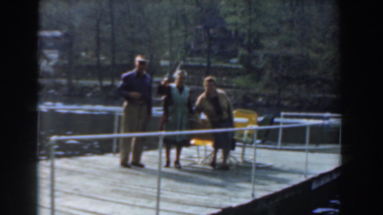 Three adults remain on the dock with chairs, railing, water, and trees behind them.
