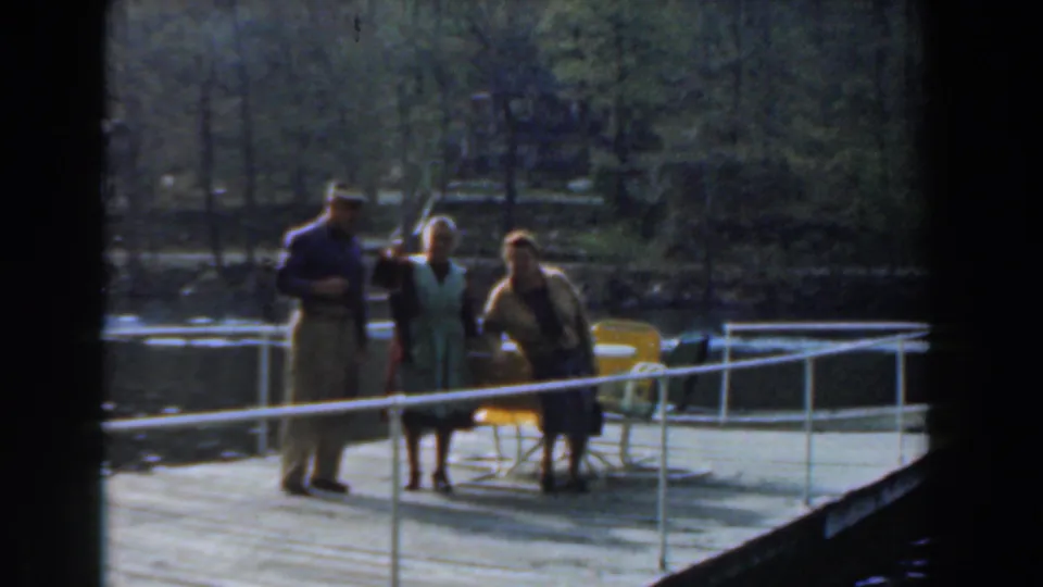 Three adults pose on the wooden dock in 1961 Novi, Michigan footage, with a railing, lake water, and trees in soft 8mm color.