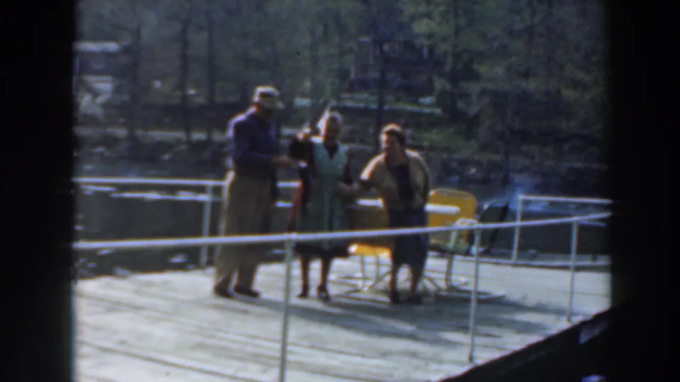 A man in a cap and two women pose on a wooden pier in 1961 home-movie footage, with one person holding a fish toward the camera.
