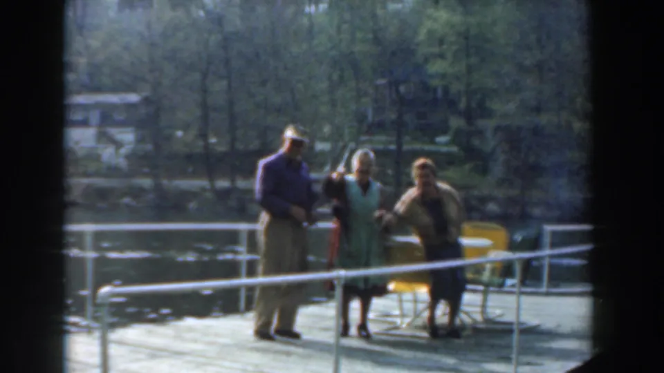 The 1961 home-movie frame shows three adults on a Novi dock beside patio chairs, with wooded lakefront visible behind them.