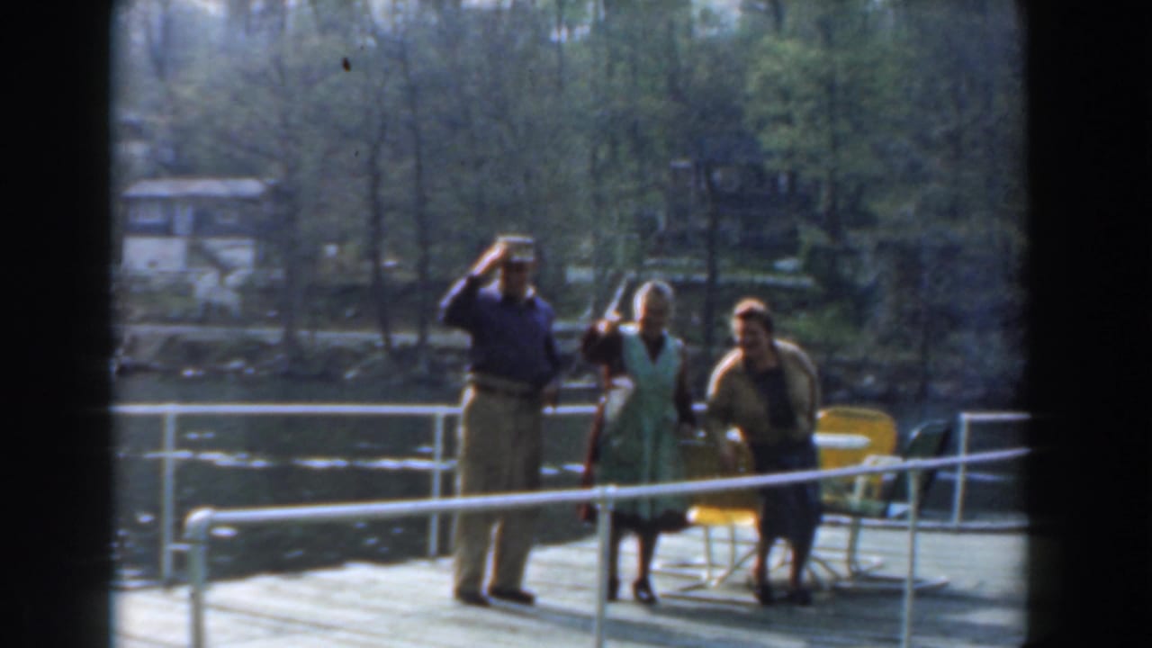 Three adults stand on a pier by the water, with one man raising his hand toward the camera.