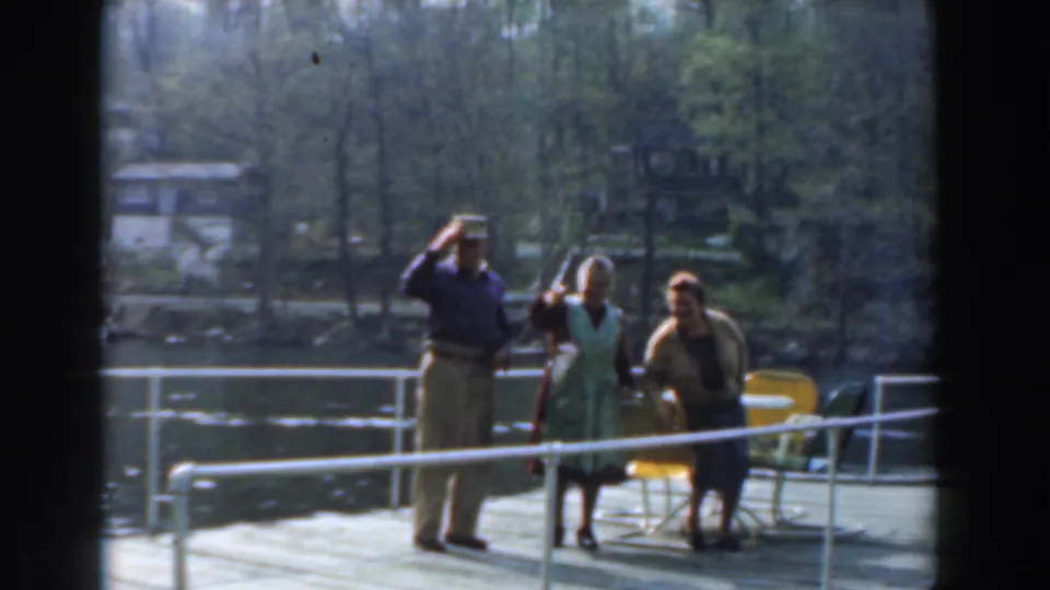 In 1961 Novi, Michigan home-movie footage, three adults pose on a lakeside pier as the man waves and the women face the camera.
