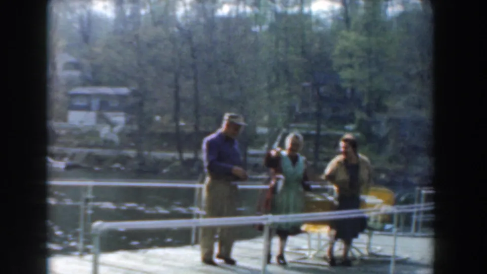 Three adults stand together on a Novi lakeside pier, with yellow chairs behind them and trees across the water.