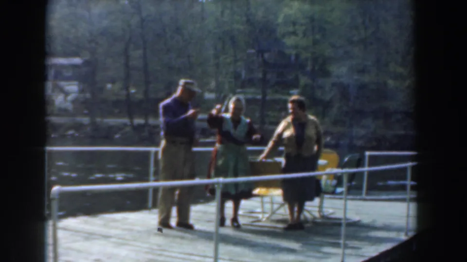 Adults on a 1961 Novi pier continue posing by the lake, with soft 8mm color and trees in the background.