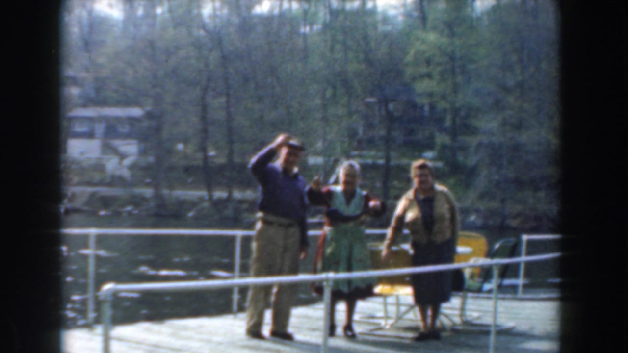 In 1961 Novi, Michigan home-movie footage, three older adults stand on a dock by lake water with trees and houses beyond.