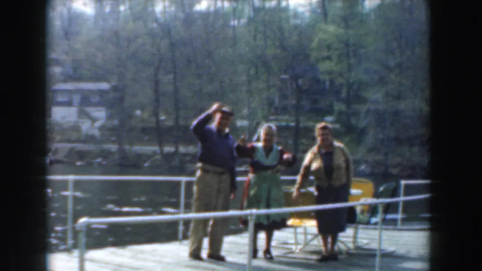 In 1961 Novi, Michigan home-movie footage, three older adults stand on a dock by lake water with trees and houses beyond.