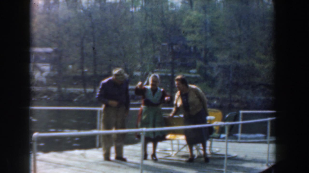 An older man and two women stand near folding chairs on a dock by the water.