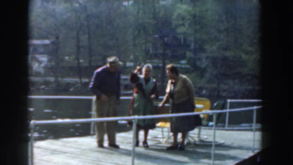 A candid 1961 Novi lakeside moment shows three older adults near a white dock railing, chairs, water, and trees.