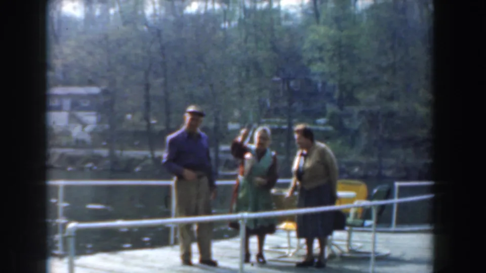 The 1961 Novi home movie frames three older adults beside yellow chairs on a dock, with lake water and wooded homes in the background.