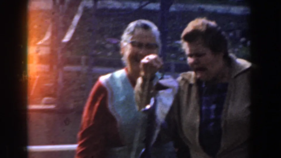 Close home-movie view of two women in 1961 Novi, Michigan, with one lifting a fish as both stand near the lakeside dock railing.