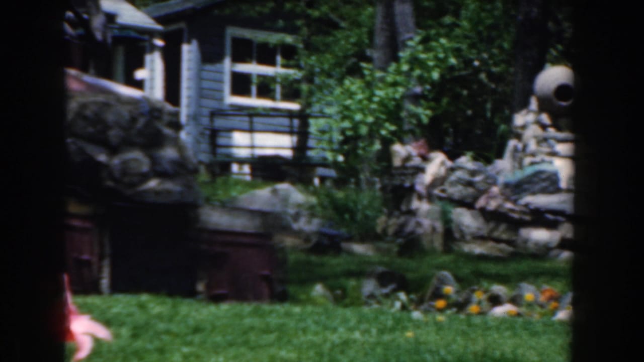 Garden lawn with flowers, trees, stone structure, and house exterior in the background.
