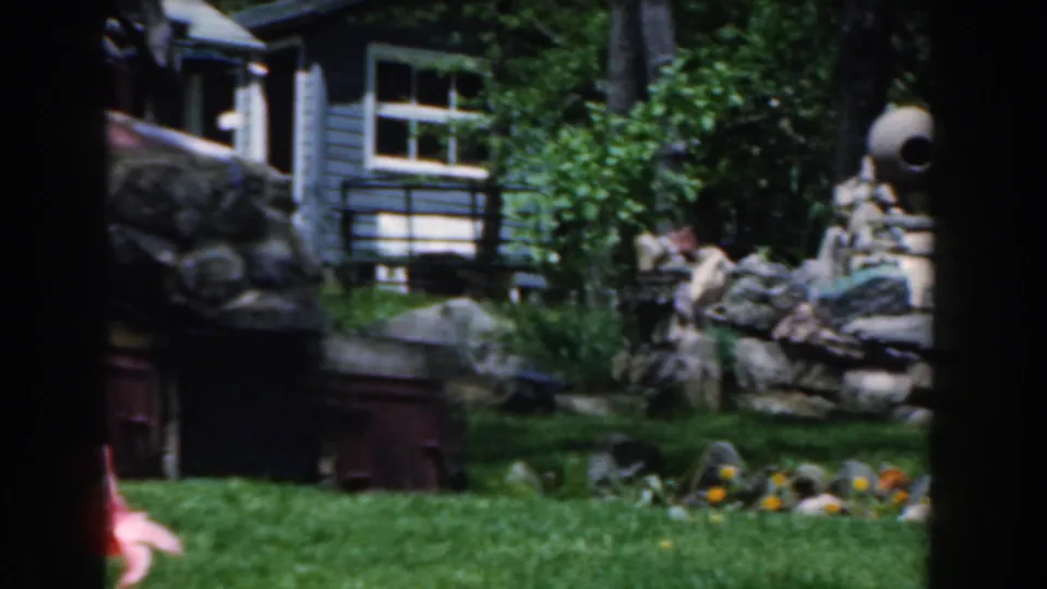 1961 Novi, Michigan home-movie frame showing a green lawn, garden flowers, trees, stone yard structure, and a house beyond.