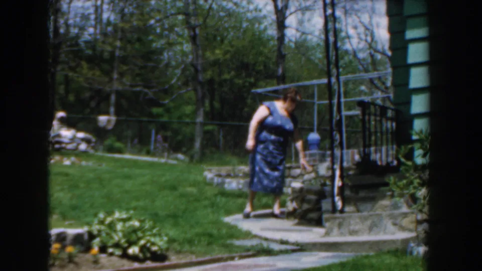 1961 Novi, Michigan backyard scene with a woman in a blue patterned dress near steps, railing, green lawn, trees, and house siding.