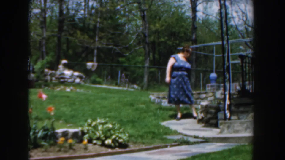 1961 Novi, Michigan frame showing the woman moving along a backyard stone path with lawn, tulips, trees, and garden structures behind her.