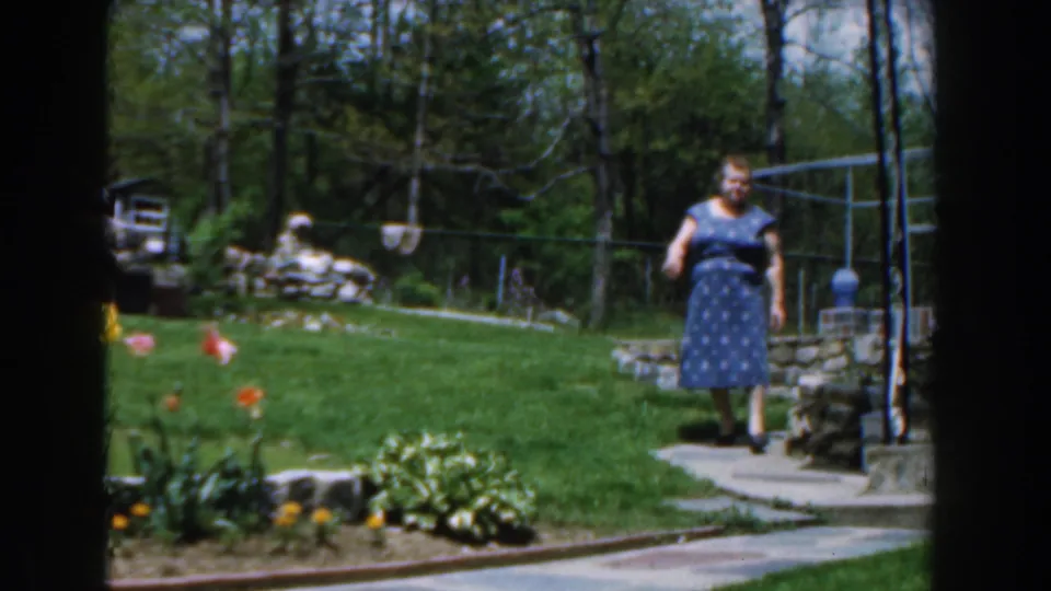1961 Novi, Michigan home-movie frame of the woman facing forward on the garden path, with tulips, lawn, stone edging, and trees visible.