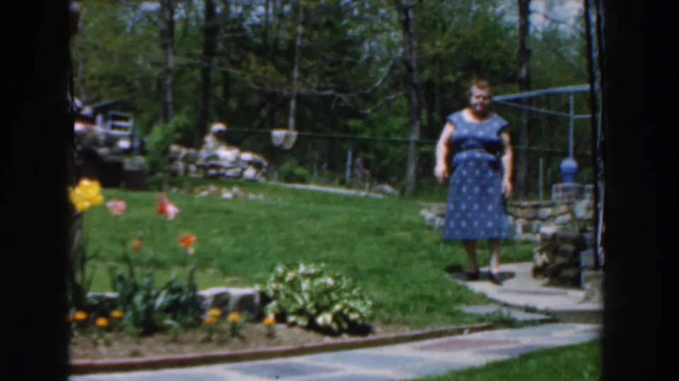 1961 Novi, Michigan frame of a woman walking toward the camera along a curved backyard path, bordered by flowers, grass, trees, and stonework.