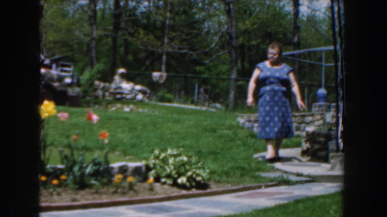 1961 Novi, Michigan home-movie frame showing a woman in a blue patterned dress walking through a sunny backyard garden.