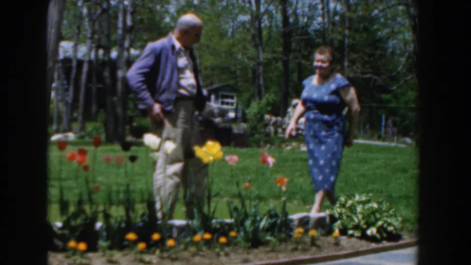 1961 Novi home-movie frame of a couple standing together beside tulips and low garden borders in a wooded backyard.