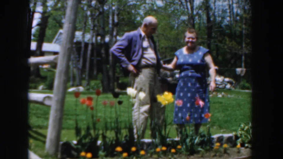 1961 Novi, Michigan frame showing an older man and woman conversing near tulips, trees, lawn, and a garden walkway.