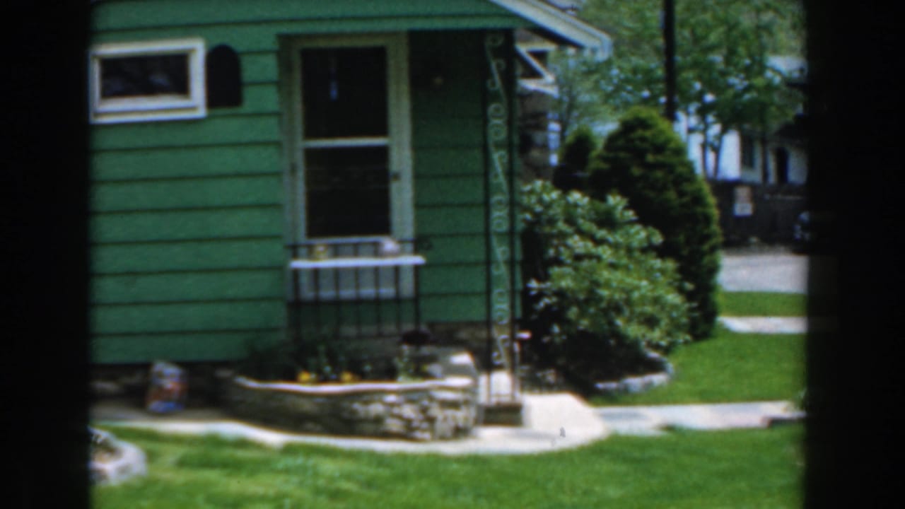 Green house exterior with front door, window, shrubs, lawn, and walkway.