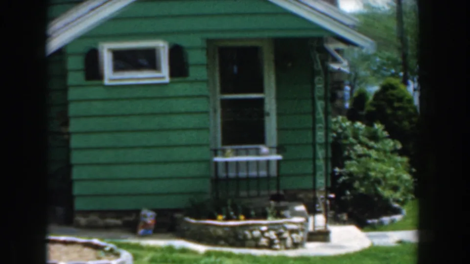 1961 home-movie view of a green Novi house exterior, showing the front window, door, planter edge, lawn, and shrubs.