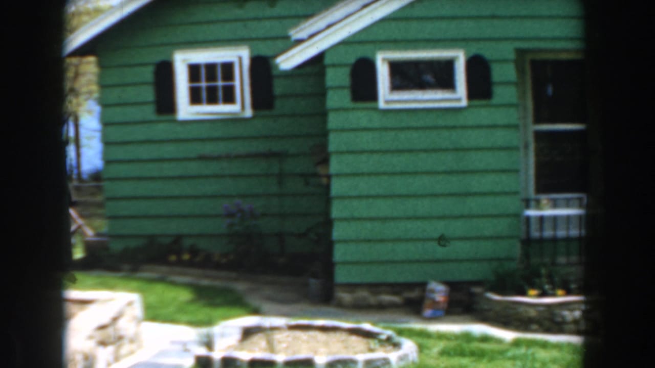 Green house exterior with white-trimmed windows, black shutters, lawn, and stone garden edging.