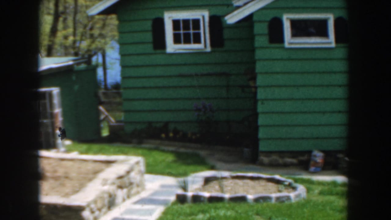 Green house facade with small windows, black shutters, stone wall, path, lawn, and trees.