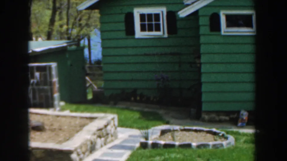 1961 Novi, Michigan 8mm frame showing the green house side, a nearby green structure, stonework, lawn, and garden edging.