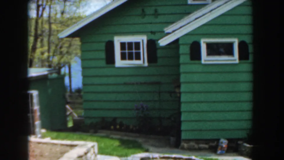 1961 Novi, Michigan frame of a green-sided residential exterior with white windows, black shutters, lawn, trees, and stone garden border.