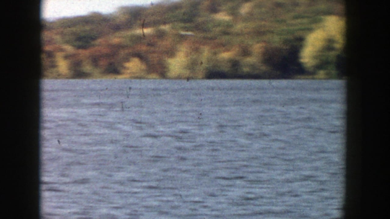 1957 Wisconsin lake scene showing rippled water, autumn-colored hillside, and dark edge framing from the filming position.