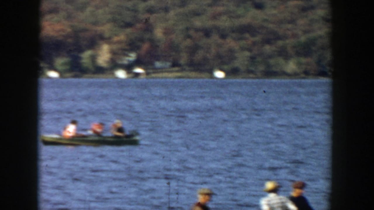 1957 Wisconsin 8mm frame with a small canoe carrying distant paddlers across open water near an autumn wooded shore.