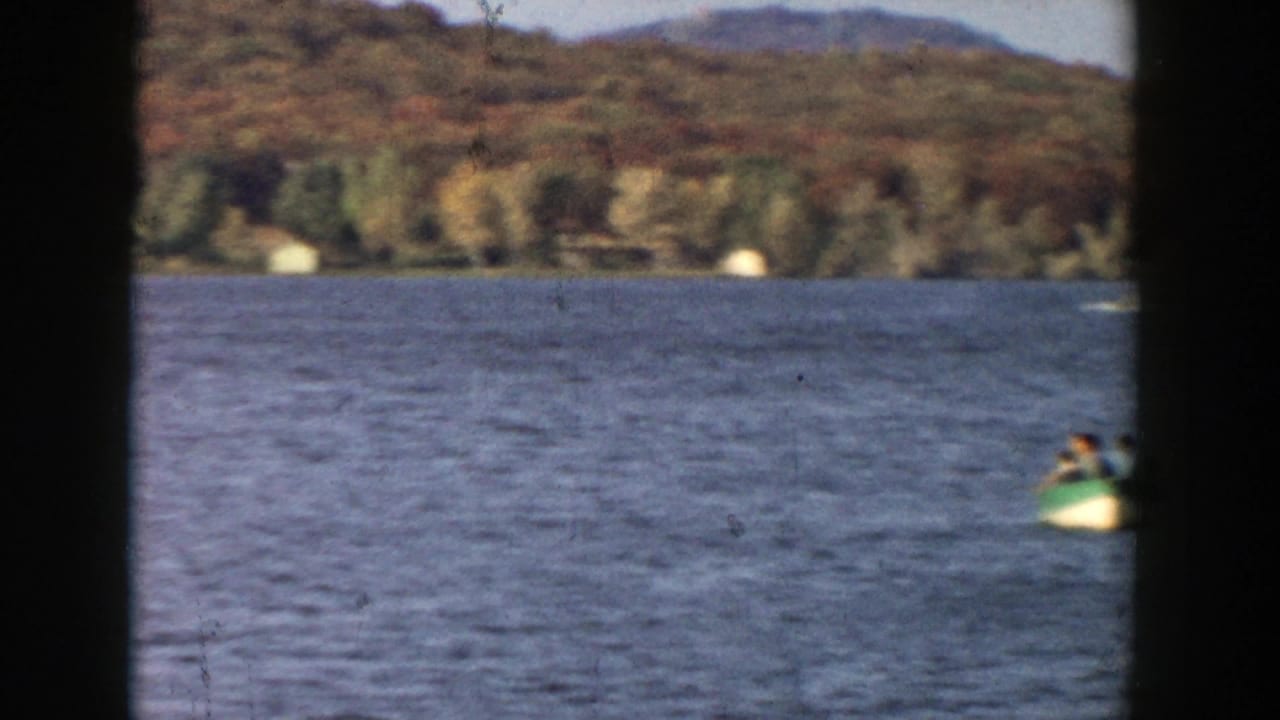 1957 Wisconsin lake view with a small paddle boat at the right edge, distant wooded hills, and broad open water.