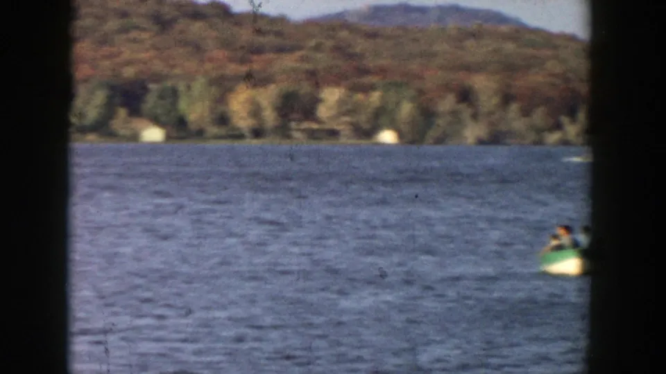 1957 Wisconsin lake view with a small paddle boat at the right edge, distant wooded hills, and broad open water.