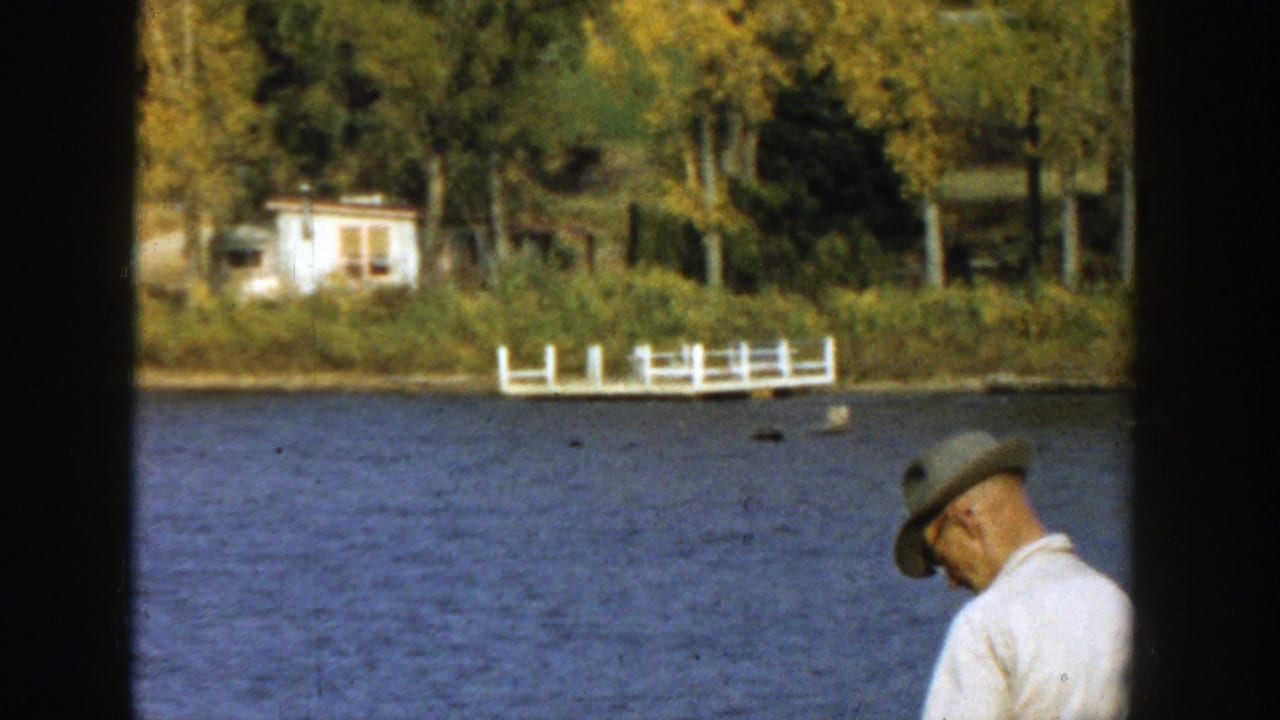 Man in a light shirt and cap stands by a lake with autumn trees and a white dock across the water.