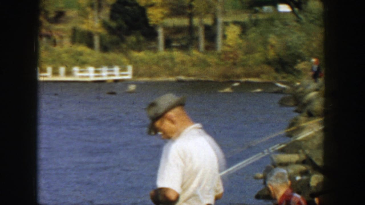 Man in a cap stands near fishing rods by the lake, with rocks and trees along the shoreline.