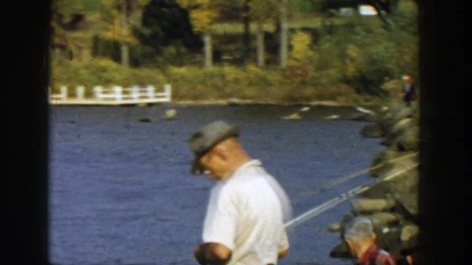 1957 Wisconsin lakeside fishing view with a capped man in the foreground, rods angled over the water, rocks, trees, and a distant white dock.