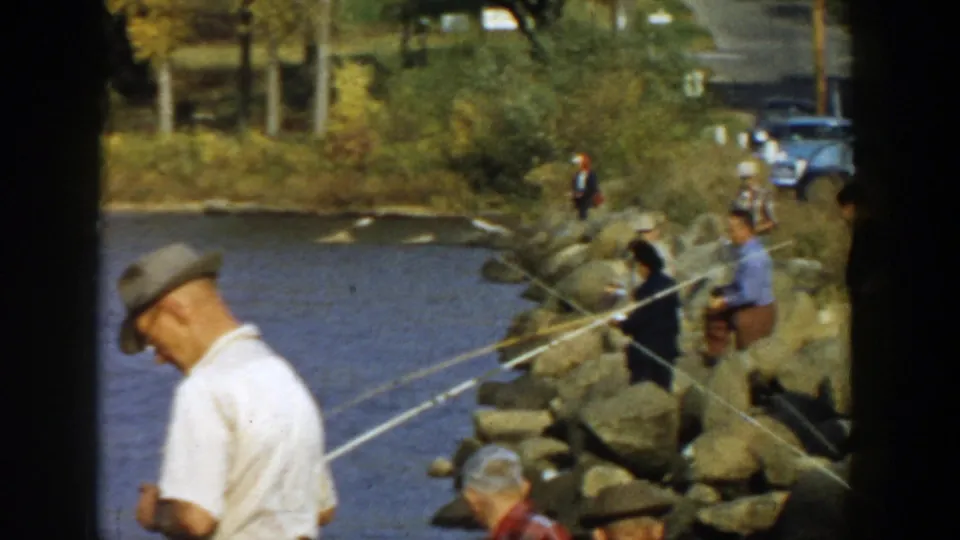 1957 Wisconsin home-movie frame with anglers on a rocky lakeshore, fishing rods over the water, and parked midcentury cars near a road behind them.