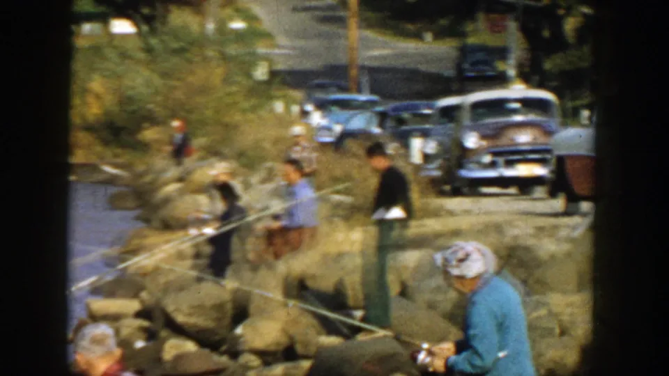 1957 Wisconsin lakeshore scene with several adults fishing on rocks, a person in a blue top in front, and classic cars parked along the road.