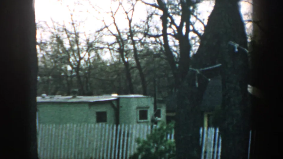 1957 Wisconsin 8mm frame showing a white picket fence, modest trailer, brick shed, and leafless wooded surroundings.