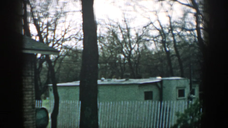 1957 Wisconsin home-movie frame of a trailer behind a white picket fence, set among bare trees under an overcast sky.