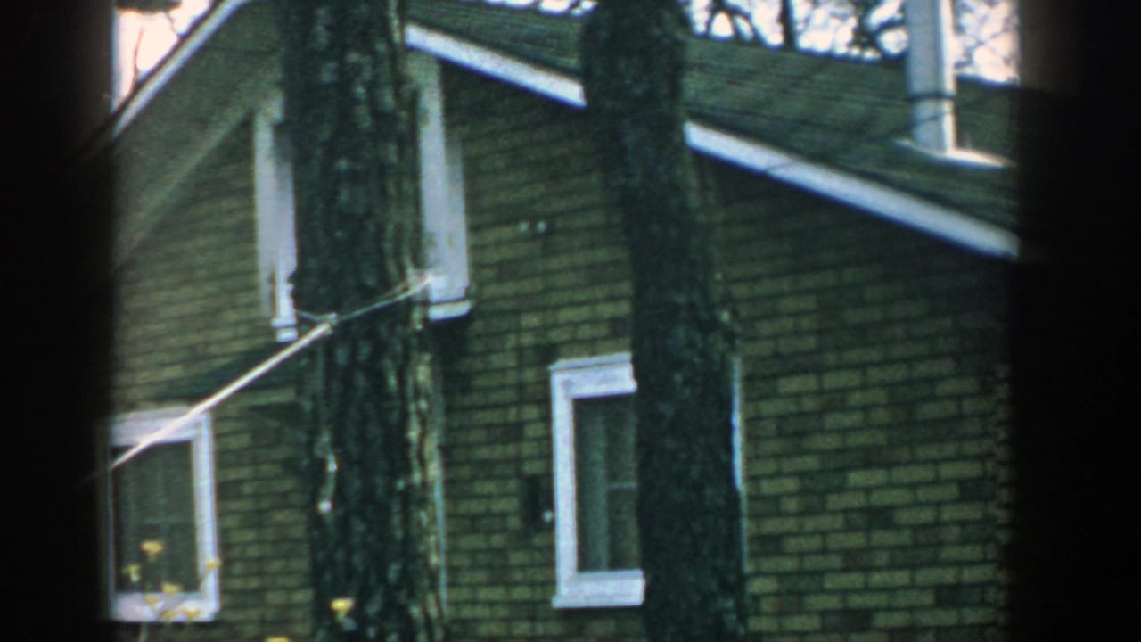 1957 Wisconsin 8mm frame showing a brick house exterior behind large tree trunks, with white-trimmed windows and sparse branches.
