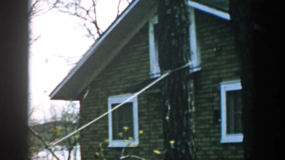 1957 Wisconsin home-movie view of a brick house gable and white-trimmed windows, partly obscured by trees and a diagonal line.